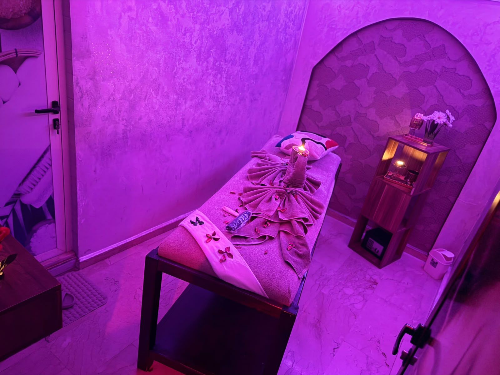 Vue plongeante d’une cabine de soin à l’arche orientale, table habillée de serviettes et pétales sous lumière rose
