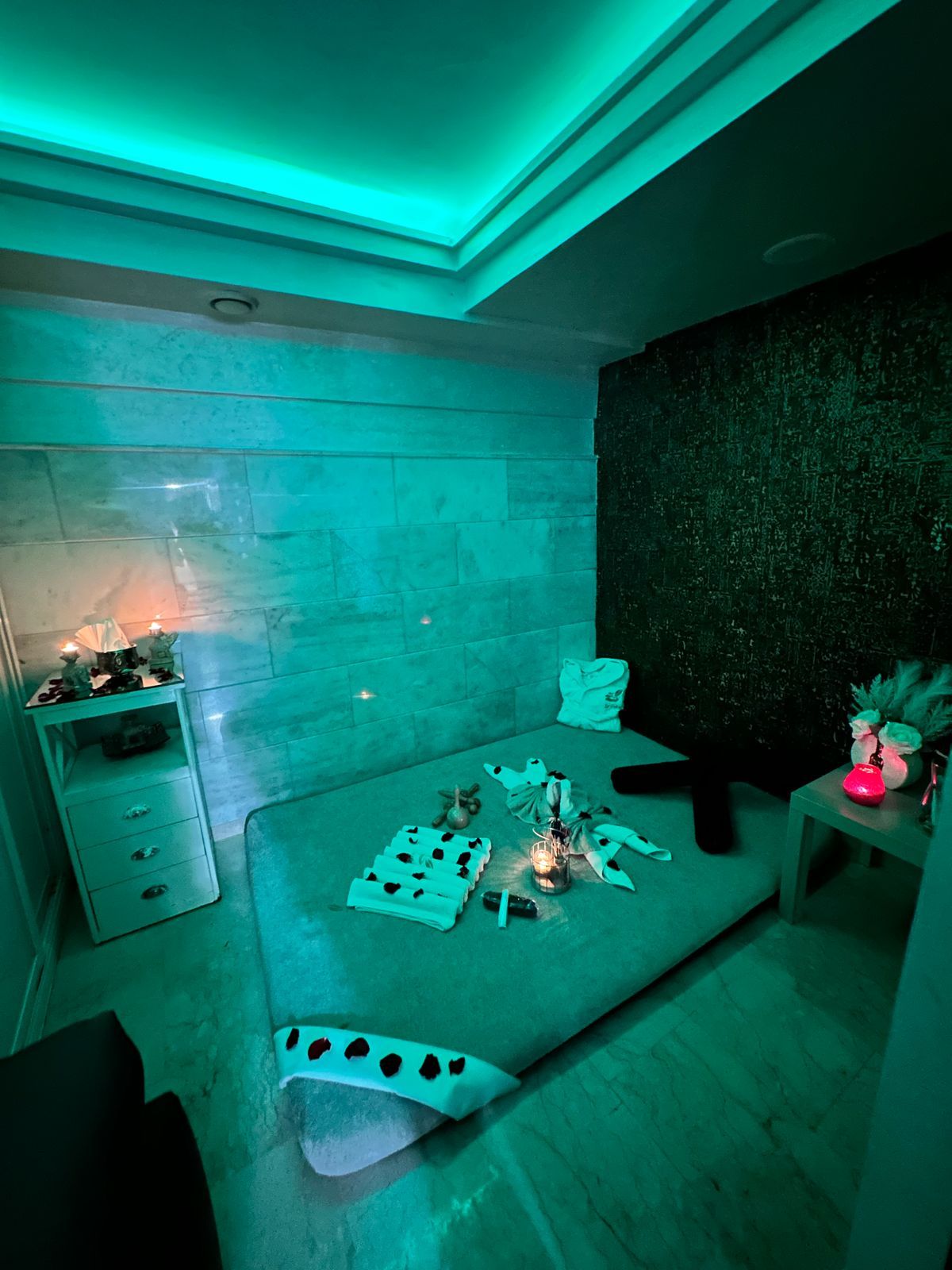 Espace duo de Le Cèdre Lounge Spa avec large matelas de massage, serviettes et bougies sous une lumière émeraude