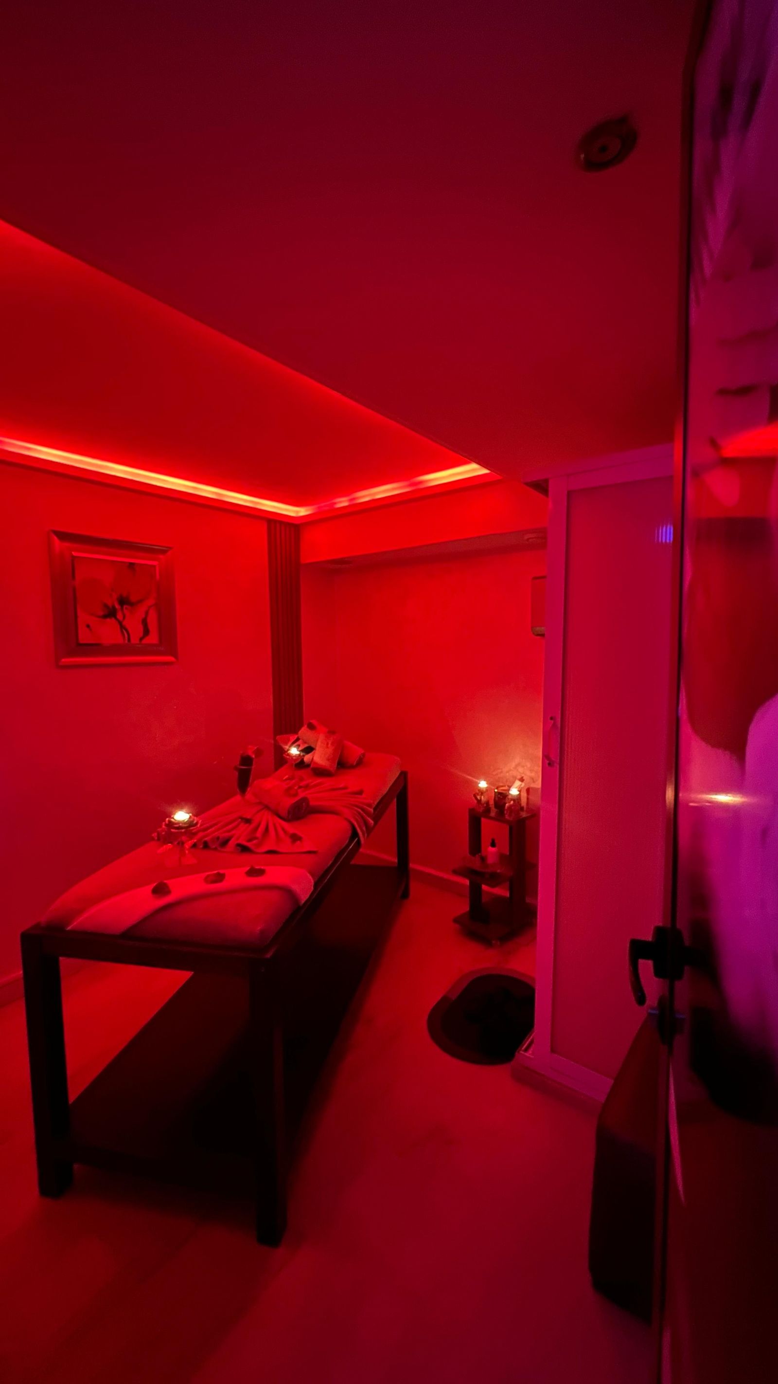 Cabine de massage privative baignée de lumière rouge, table habillée de serviettes et pétales, bougies allumées