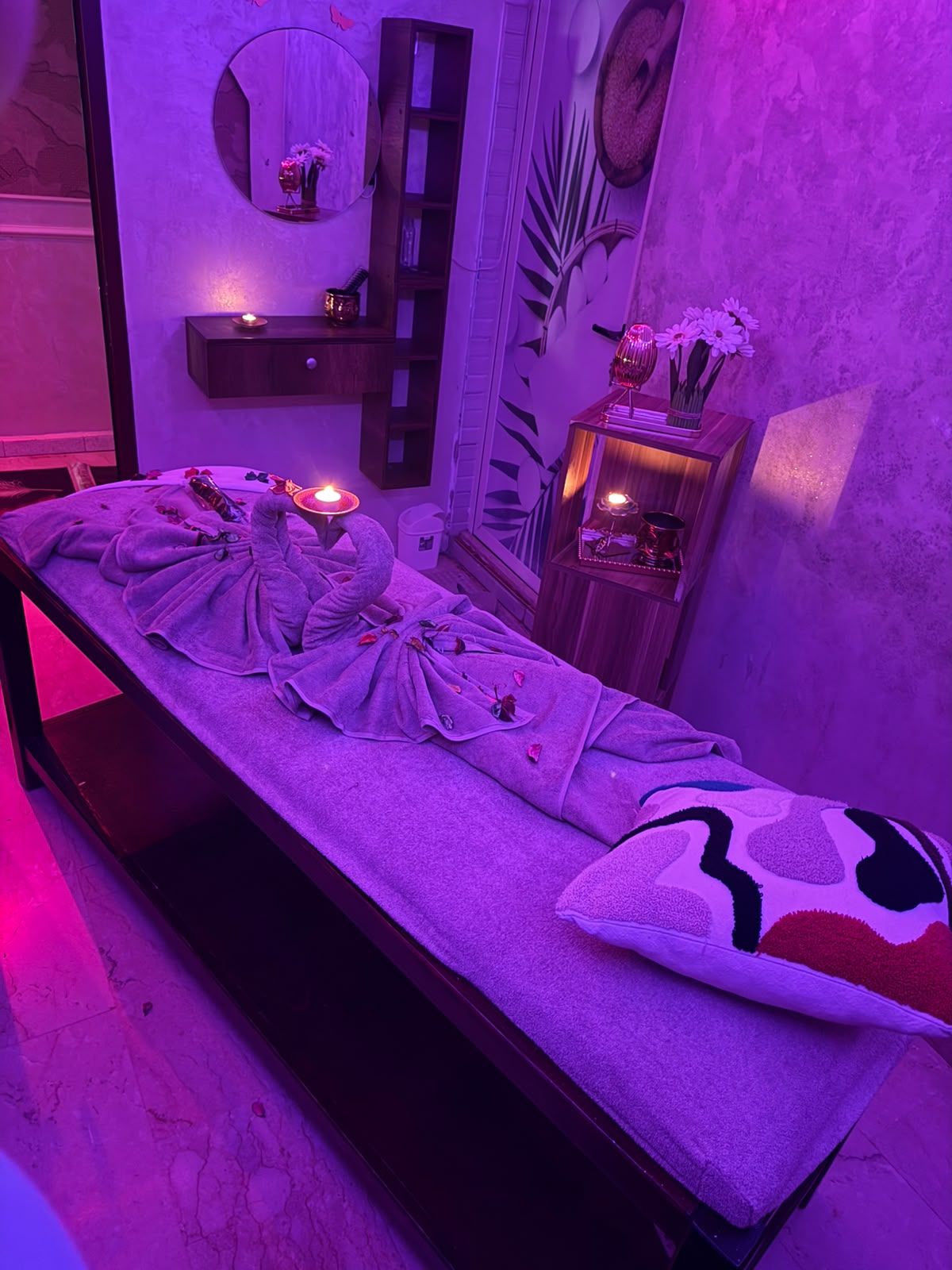 Cabine de massage de Le Cèdre Lounge Spa sous lumière violette avec miroir rond, étagères en bois et serviettes pliées en cygne