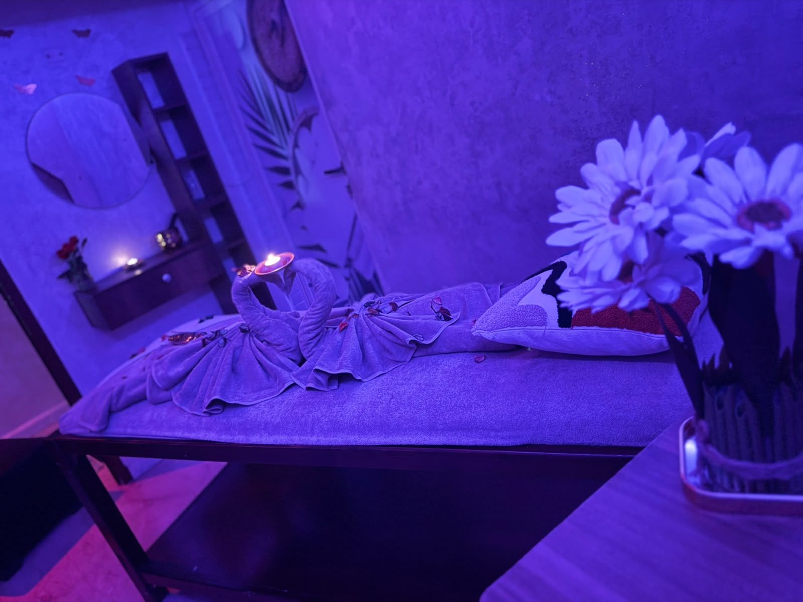 Cabine de soin de Le Cèdre Lounge Spa sous lumière violette : table de massage avec serviettes pliées en cygnes, fleurs blanches et bougie