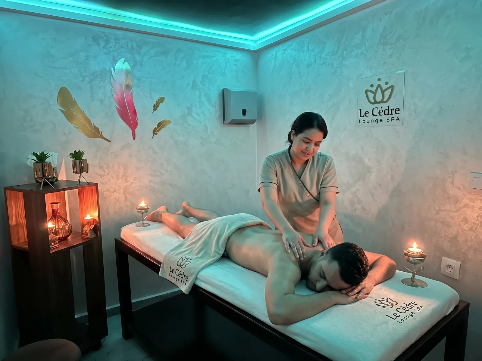 تدليك مريح للظهر في مقصورة خاصة في Le Cèdre Lounge Spa بالدار البيضاء، سقف بإضاءة فيروزية وريش زخرفي على الجدار وشموع